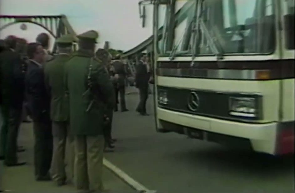 A video frame on a bridge deck under a green steel truss: a cream coach stands beside a line of uniformed guards in green with peaked caps and men in dark suits
