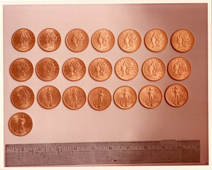 Twenty-two gold twenty-dollar coins laid out in three rows of seven and one below on a pinkish grey card, Liberty heads and standing Liberty figures with dates from 1897 to 1924, a wooden ruler along the bottom