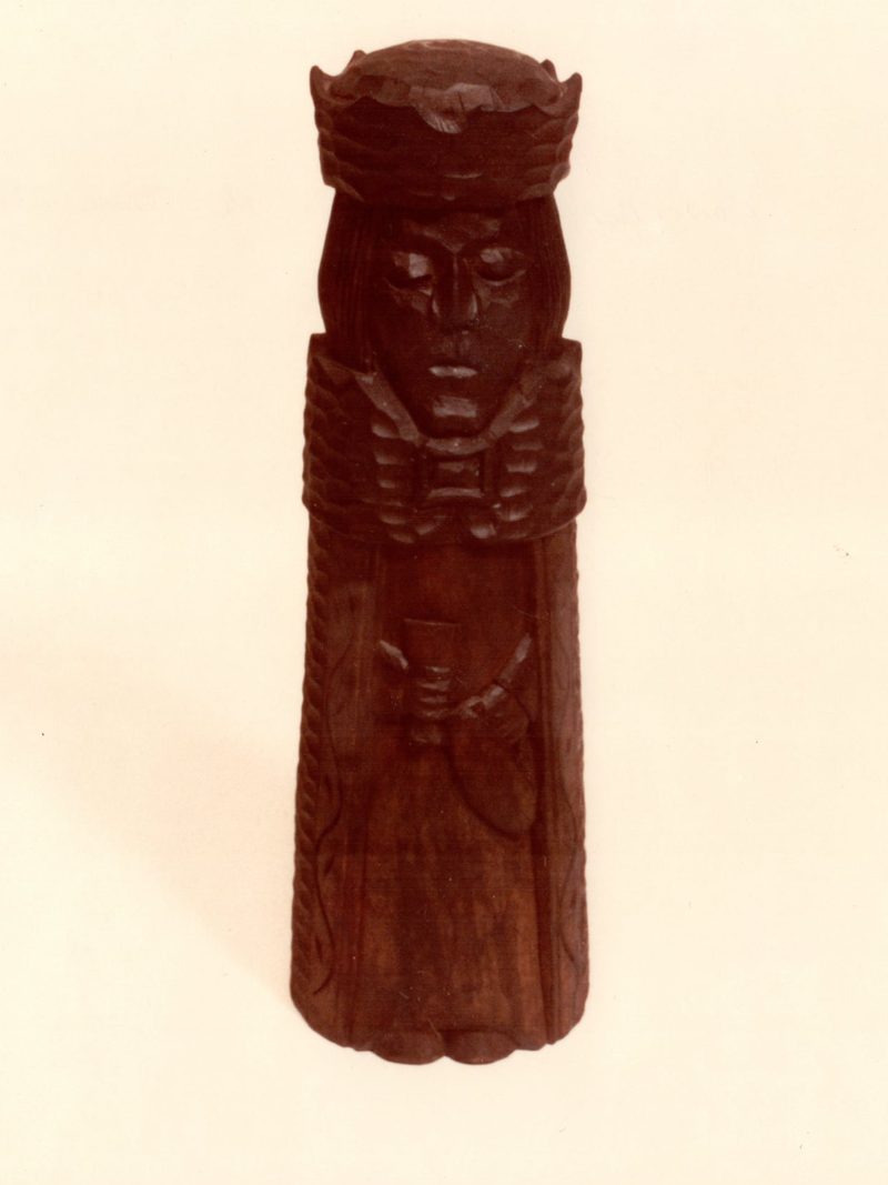 A tall carved figure of dark stained wood, crowned and robed, holding a small cup in both hands, photographed standing on cream photographic paper
