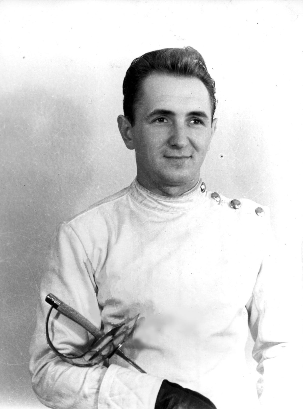 A black and white studio portrait of a smiling dark haired man in a white fencing jacket, a sabre held across his body