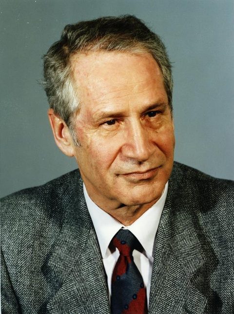 A colour studio portrait of a grey haired man in a herringbone jacket, white shirt and dark red tie, looking to one side