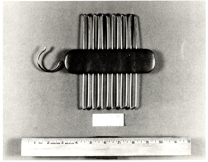 A black and white evidence photograph of a padded dark leather bar carrying a row of wire tie hooks and a wire hanging hook, a small typed evidence card below it and a twelve inch wooden ruler along the bottom