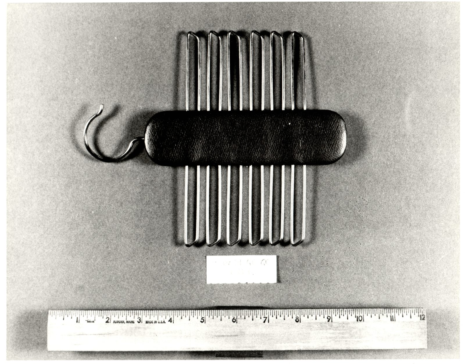 A black and white evidence photograph of a padded dark leather bar carrying a row of wire tie hooks and a wire hanging hook, a small typed evidence card below it and a twelve inch wooden ruler along the bottom