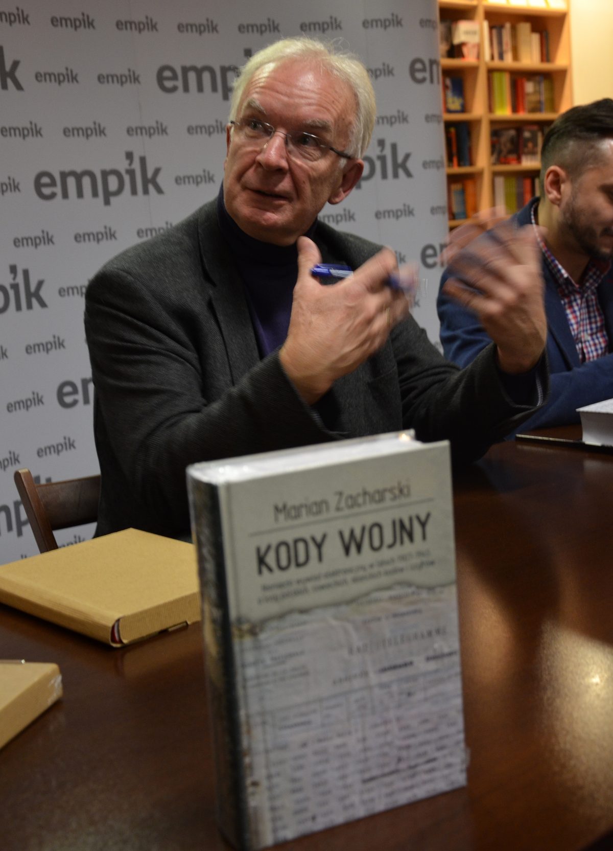 Marian Zacharski, white haired and in glasses, pen in hand, talking at a signing table in a bookshop, a copy of his book Kody wojny standing in front of him and a second man at the right edge