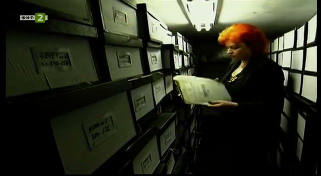 A narrow archive corridor lined with labelled boxes, a woman lifting a bundle of papers from one of the shelves
