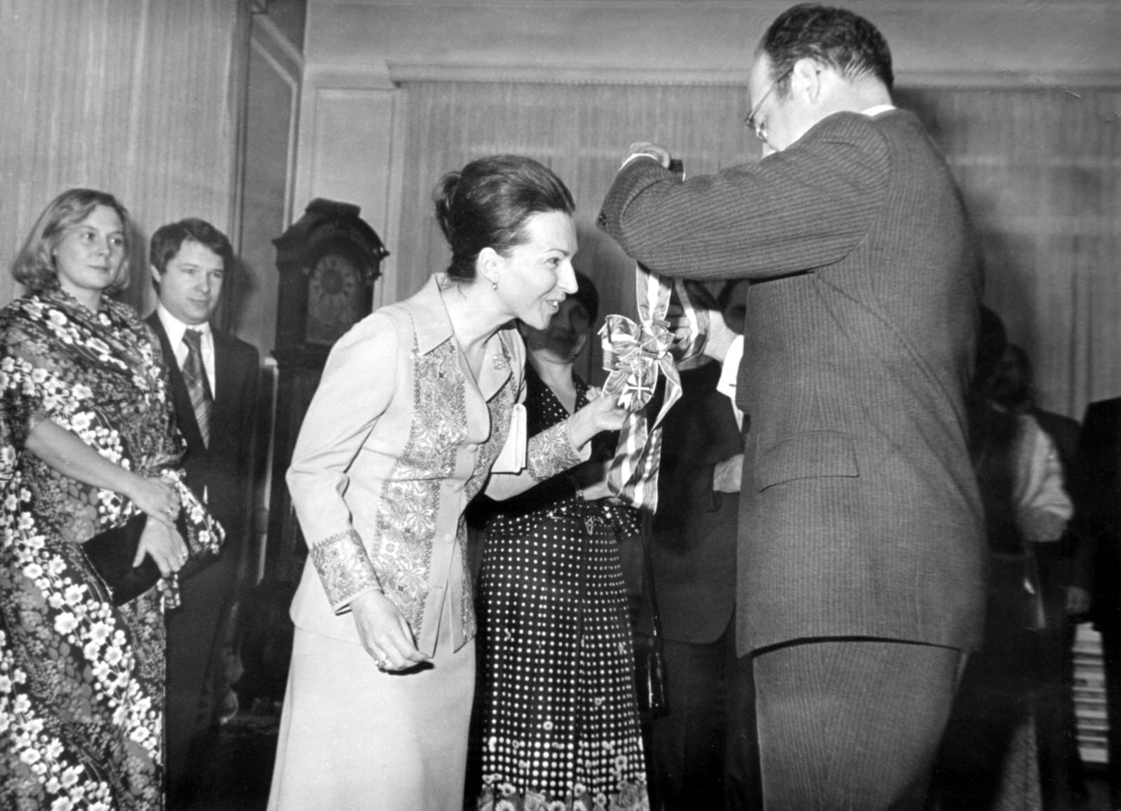 A black and white photograph of a ceremony in a panelled room: a man fastens a decoration on a striped ribbon while a woman in a light suit holds it and guests look on