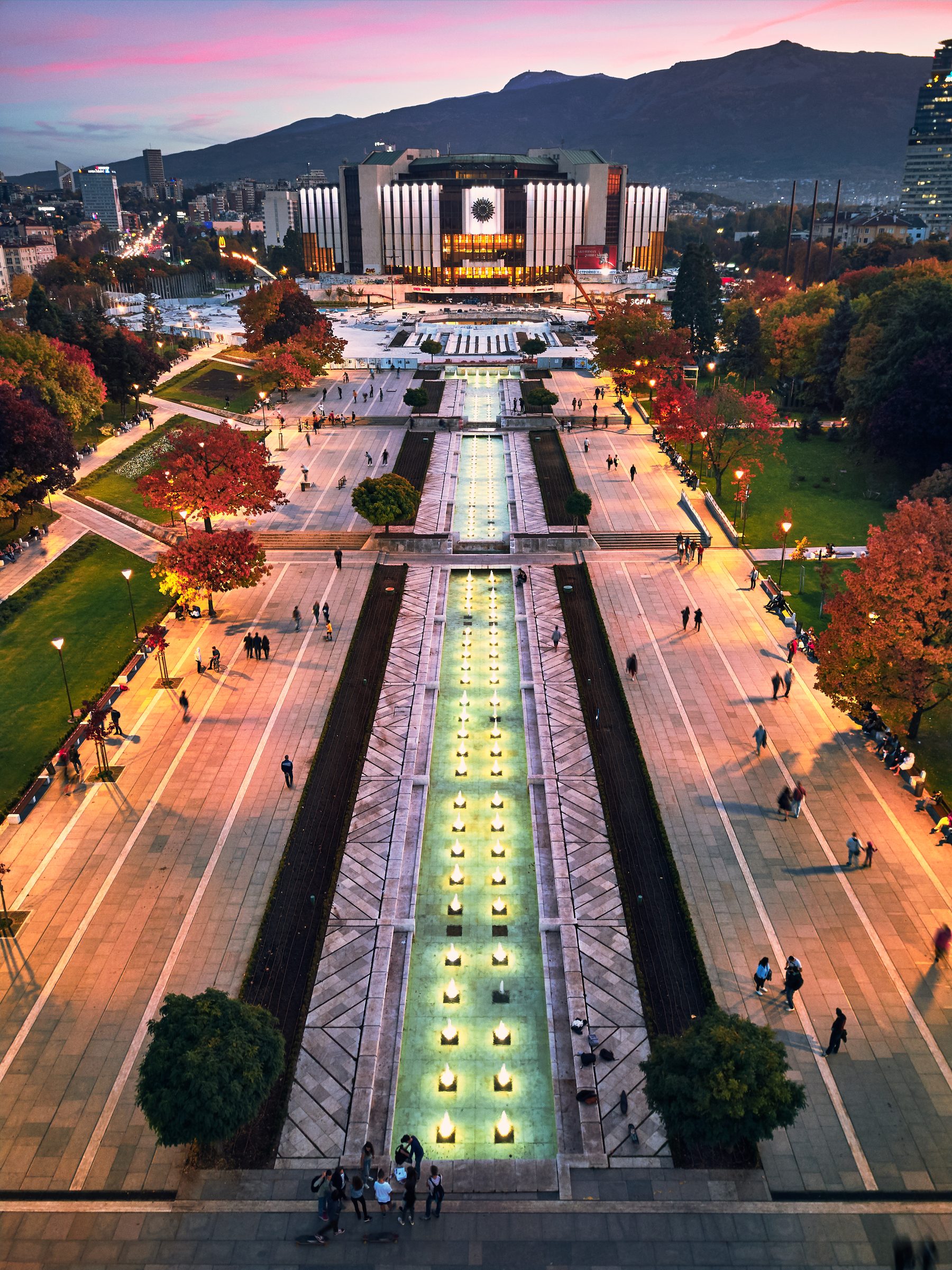 An aerial view at dusk of a large low palace of culture at the head of a long park, with lit fountains running towards it and mountains behind