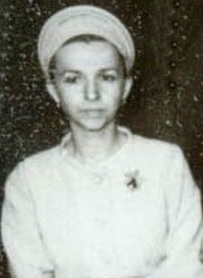 A small black and white portrait of a woman in a pale head wrap and a light jacket with a brooch, looking towards the camera