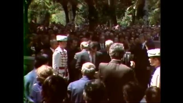 Colour film of a crowd filling a tree lined street, with uniformed guards in white tunics and plumed caps standing among the mourners