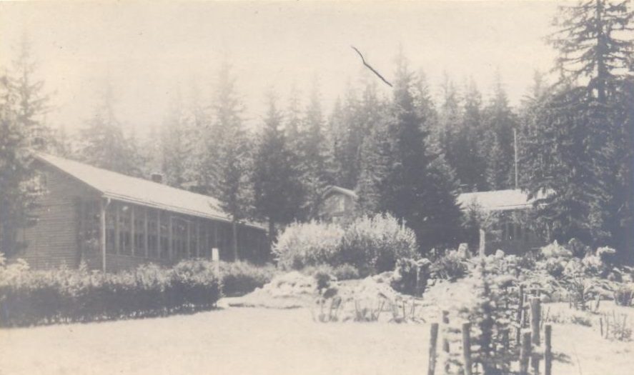 An archive photograph of a long low timber building with a glazed veranda standing in snow among tall conifers, further roofs visible through the trees