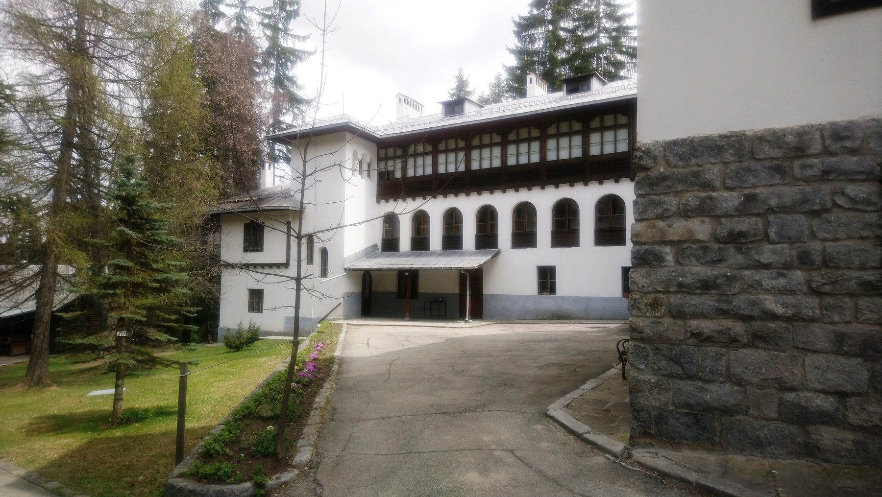 A white mountain residence of two storeys with a long glazed timber gallery above and arched windows below, standing among conifers