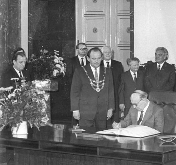 A black and white photograph of an elderly man in glasses seated at a table signing a large open book, a man wearing a chain of office standing behind him with other officials looking on